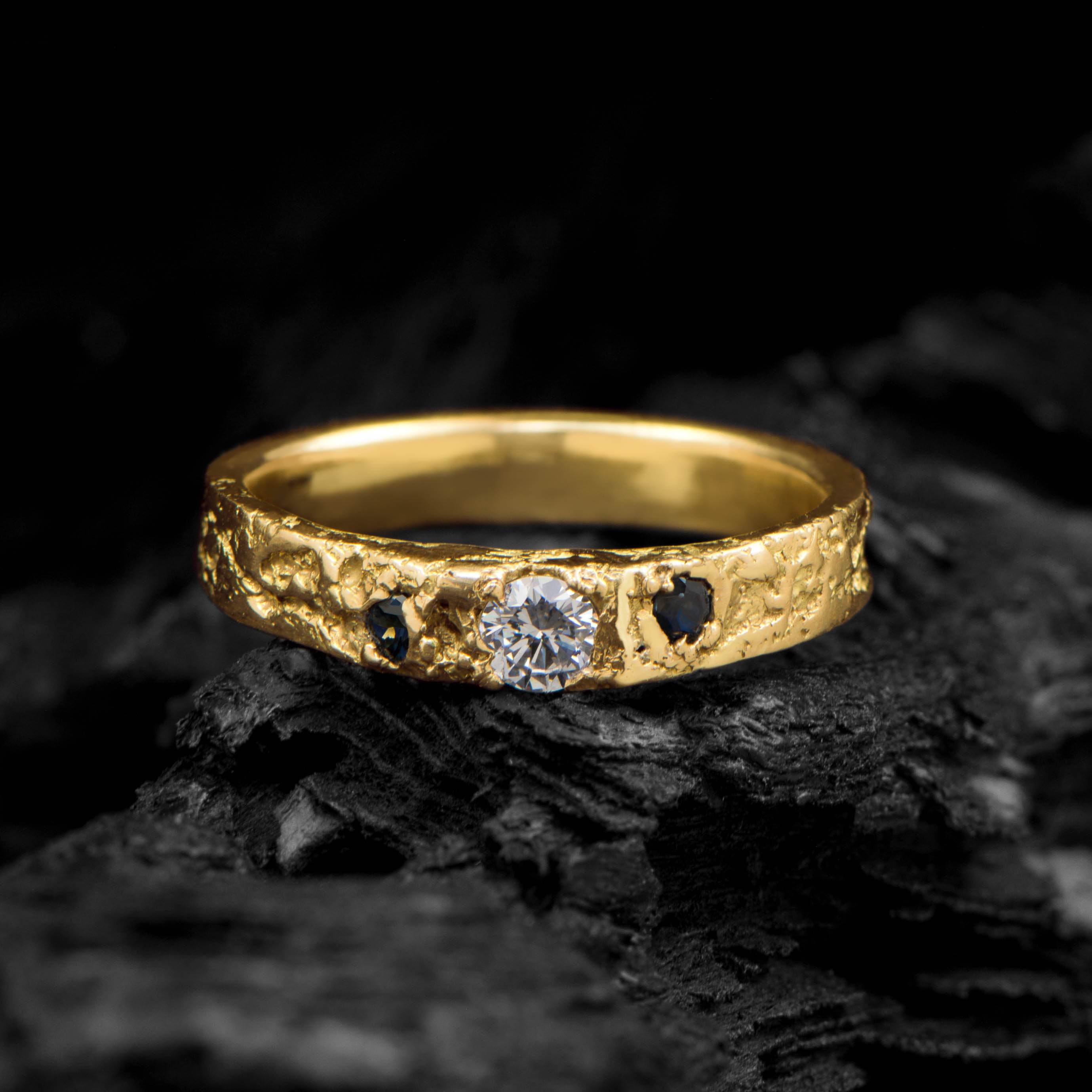 Gold ring with a diamond and two dark blue sapphires on a dark textured background