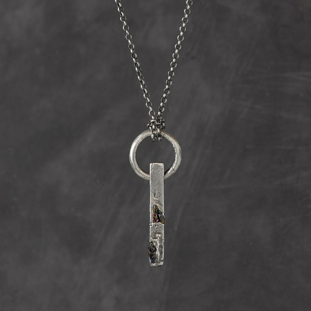 Handcrafted sterling silver necklace with sculptural bar pendant and suspended silver ring on a grey background.