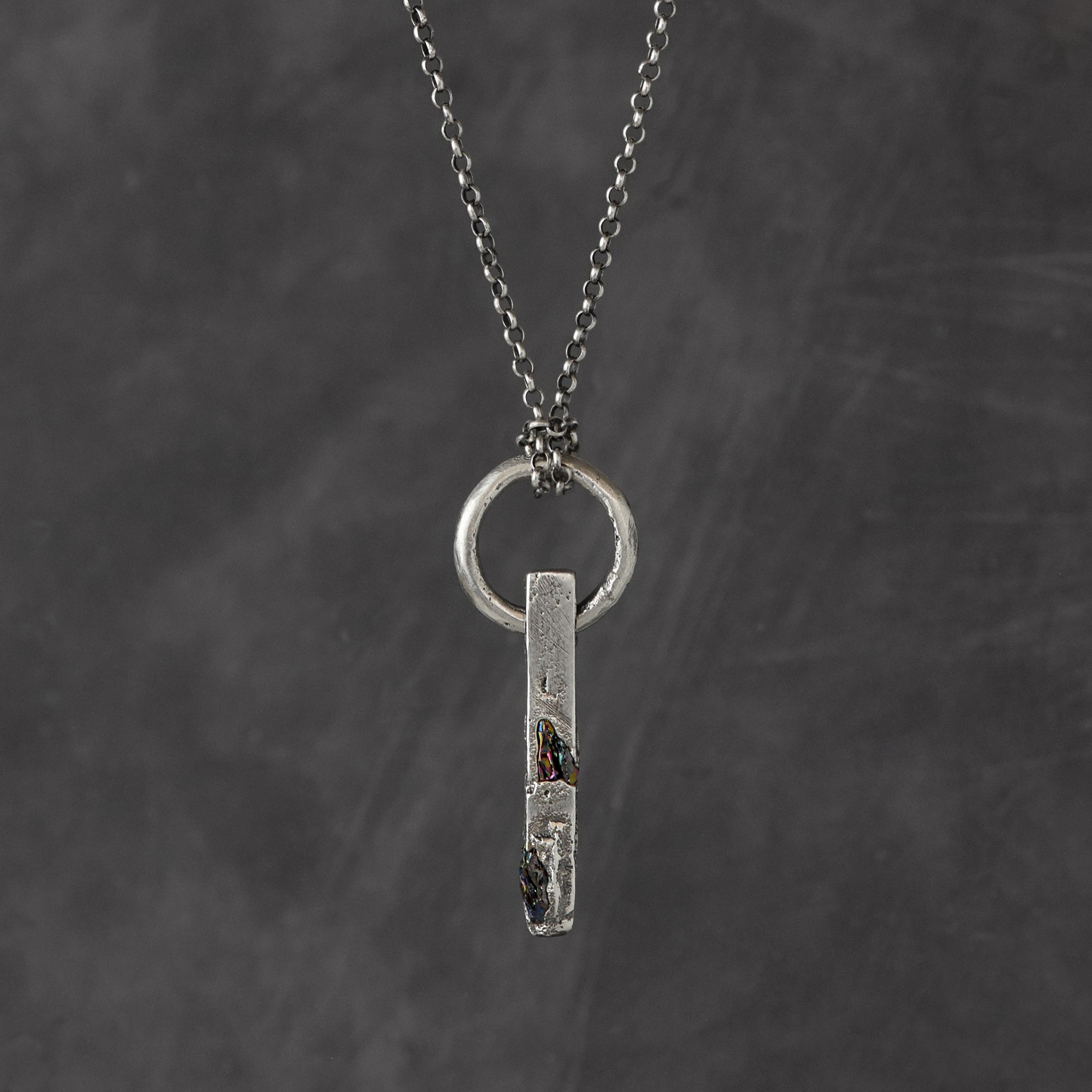 Handcrafted sterling silver necklace with sculptural bar pendant and suspended silver ring on a grey background.