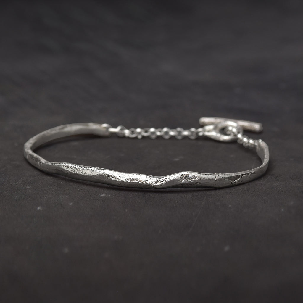 Slightly textured sterling silver cuff bracelet with a melted shape and a chain with toggle clasp on a dark background.