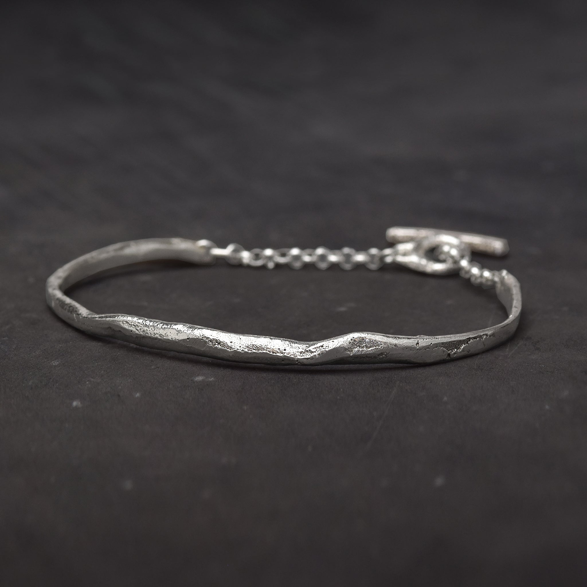 Slightly textured sterling silver cuff bracelet with a melted shape and a chain with toggle clasp on a dark background.