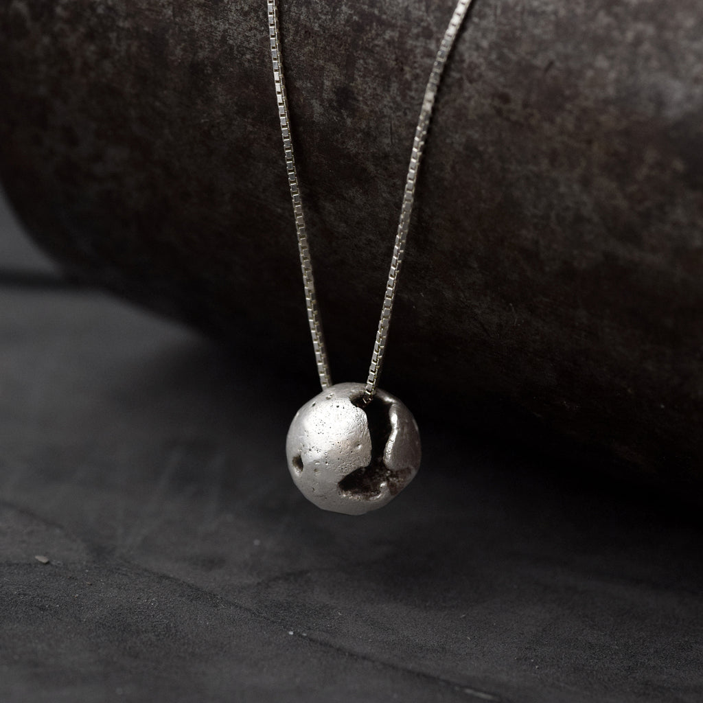 A silver pendant with a spherical shape with craters and a rugged texture suspended from a silver curb chain in the jewellers workshop.
